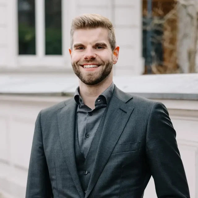 Julian Schwarz im dunklen Anzug steht vor einem hellen Gebäude und blickt lächelnd in die Kamera.